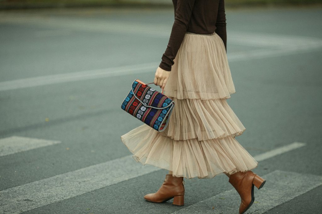 Woman in elegant skirt and boots crossing a zebra crossing, holding a boho-style bag.